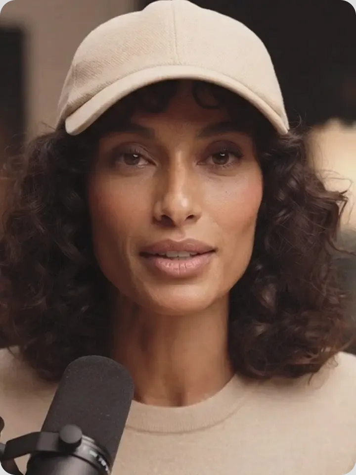A person with shoulder-length dark curly hair wearing a light beige baseball cap and matching crewneck shirt speaks into a black podcast microphone.